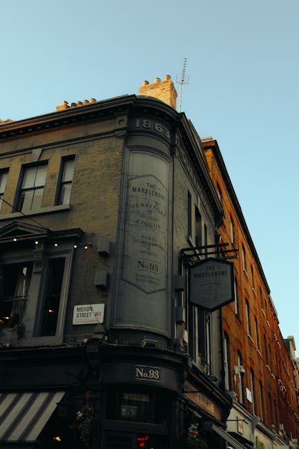 Photograph of the exterior corner of a historic building located at No. 98 Baker Street in Marylebone, featuring signage for 'The Marylebone' and possible advertisements or descriptions related to the building's history. The building's facade includes large windows on the ground floor, some with awnings, and decorative architectural details such as cornices and stonework. The upper floors are made of light-colored bricks with black window frames, and the building is topped with an ornate chimney and a small antenna. The scene is set against a clear sky, with sunlight illuminating the upper section of the building, suggesting a daytime setting. This image visually relates to house removals and local property environments handled by Marylebone Man and Van, illustrating the type of urban building involved in professional furniture transport, packing, and home relocation services, especially for properties along narrow-stair or historic areas.