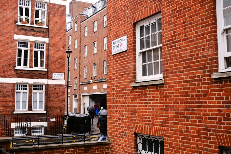The image depicts an exterior view of a red brick residential building located on Providence Court W1 in Marylebone, with two visible windows on the right side. In the foreground, part of a lower window with iron bars is seen at the bottom of the building. Two movers, dressed in casual clothing and caps, are engaged in a home relocation activity; one is carrying a large, wrapped piece of furniture, while the other is walking toward a black street-level garbage bin. The scene takes place on a pavement with a black metal fence and yellow traffic barriers, indicating a constrained urban environment. Adjacent to the building, a narrow alley leads towards other residential structures built from similar red brick materials, featuring multiple white-framed windows. The area appears well-lit with natural daylight, and signs indicating the street names are visible on the building corner and adjacent wall. The context suggests furniture transport as part of a move involving packing and loading at a Marylebone address, supported by the professional services of Marylebone Man and Van.