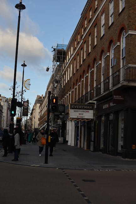 Two men are engaged in a home relocation activity outside a storefront named 'GUANG' in Marylebone, London. One man, wearing a black cap and dark clothing, holds a wheeled trolley handle and carries a