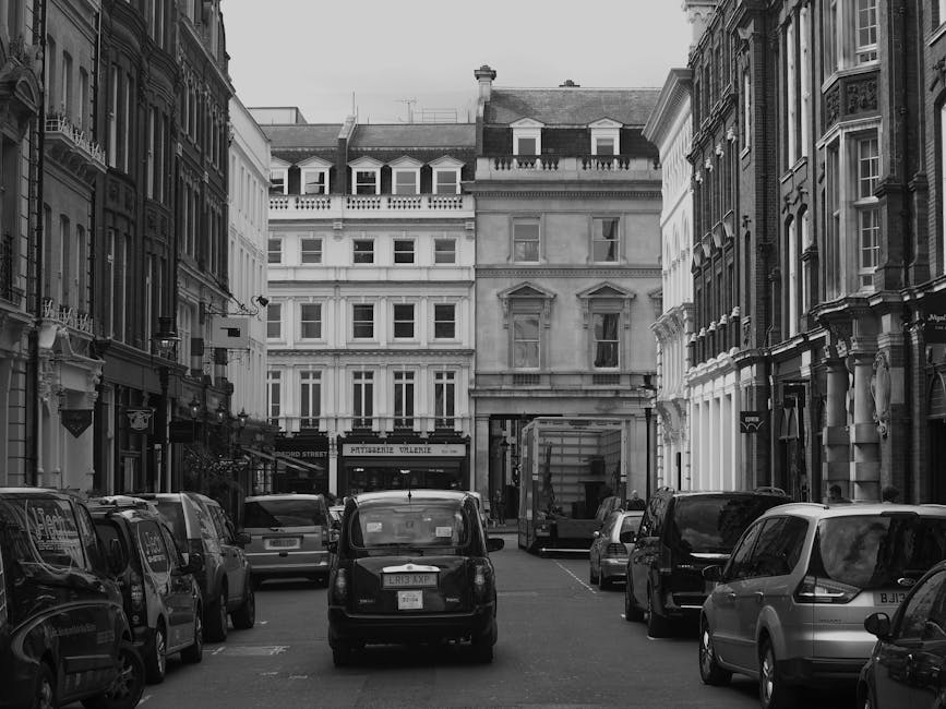 Black and white photograph of a narrow urban street with multi-storey Victorian-style buildings on both sides. Several cars, including compact vehicles and a van, are parked along the curb and on the