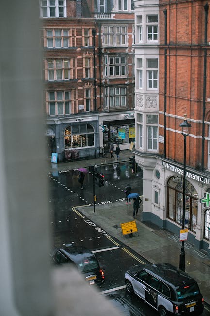 View from a high window overlooking a rainy street in Marylebone, featuring red brick Victorian-style residential buildings with bay windows and decorative detailing. The street is wet from recent rain, with pedestrians carrying umbrellas walking on the sidewalk. A black van is parked on the street, and a small yellow roadwork sign is visible near the curb. The storefront on the ground floor includes a medical centre, with signage partially visible. In the foreground, part of a white window frame or wall is visible, indicating the perspective from inside a building. This scene illustrates the urban environment where house removals and furniture transport services by Marylebone Man and Van are often coordinated during home relocation projects, requiring careful planning and execution of loading and unloading processes.