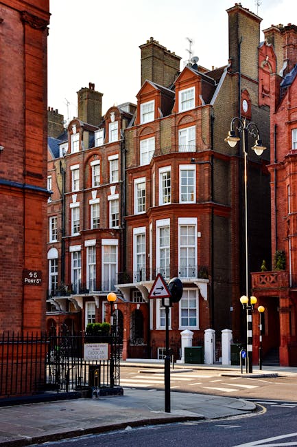 View from a high window overlooking a rainy street in Marylebone, featuring red brick Victorian-style residential buildings with bay windows and decorative detailing. The street is wet from recent rain, with pedestrians carrying umbrellas walking on the sidewalk. A black van is parked on the street, and a small yellow roadwork sign is visible near the curb. The storefront on the ground floor includes a medical centre, with signage partially visible. In the foreground, part of a white window frame or wall is visible, indicating the perspective from inside a building. This scene illustrates the urban environment where house removals and furniture transport services by Marylebone Man and Van are often coordinated during home relocation projects, requiring careful planning and execution of loading and unloading processes.